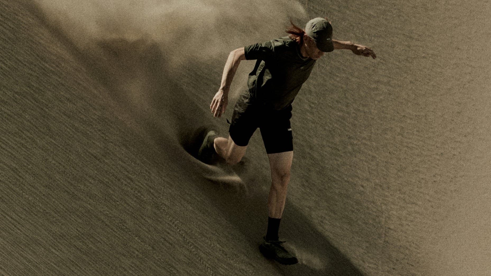 Corredor desce uma duna de areia em alta velocidade levantando poeira, com boné, camiseta e shorts escuros, em campanha da Satisfy