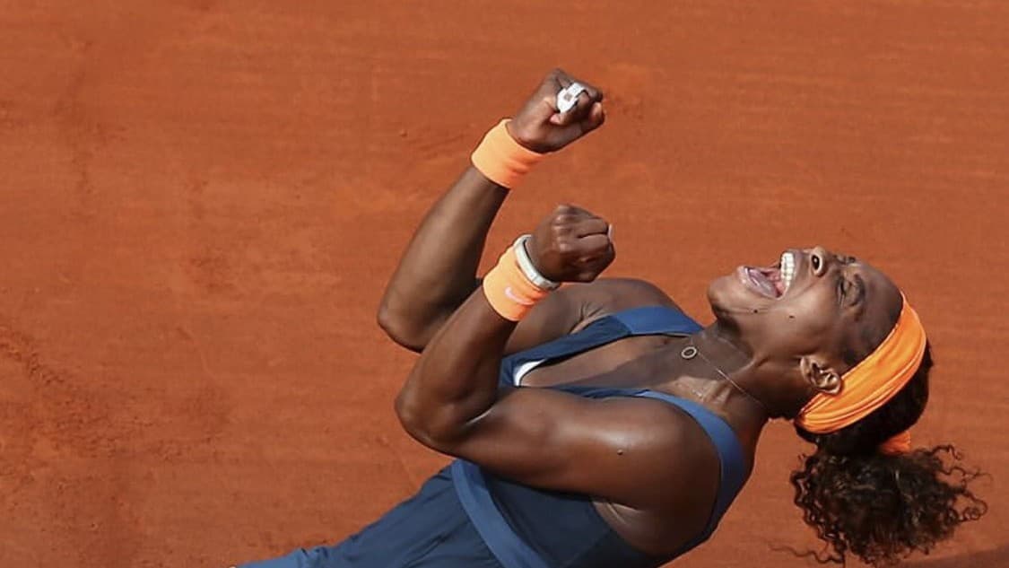 Serena Williams ajoelhada no saibro de Roland Garros, os dois punhos erguidos e a cabeça jogada para trás, comemorando a vitória