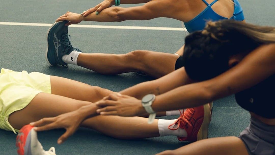 Três corredoras alongam sentadas na pista de atletismo, com tênis de corrida em destaque