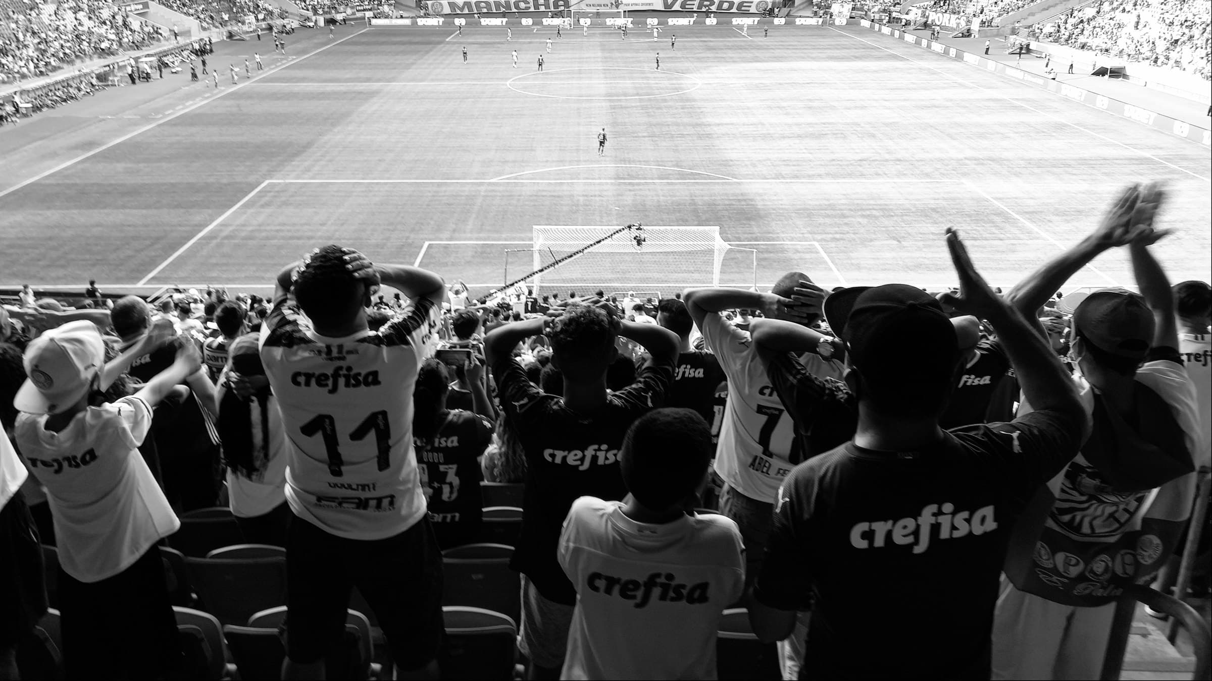 Torcida de pé na arquibancada, vista de costas, com camisas que trazem o nome do patrocinador nas costas, diante do gramado iluminado