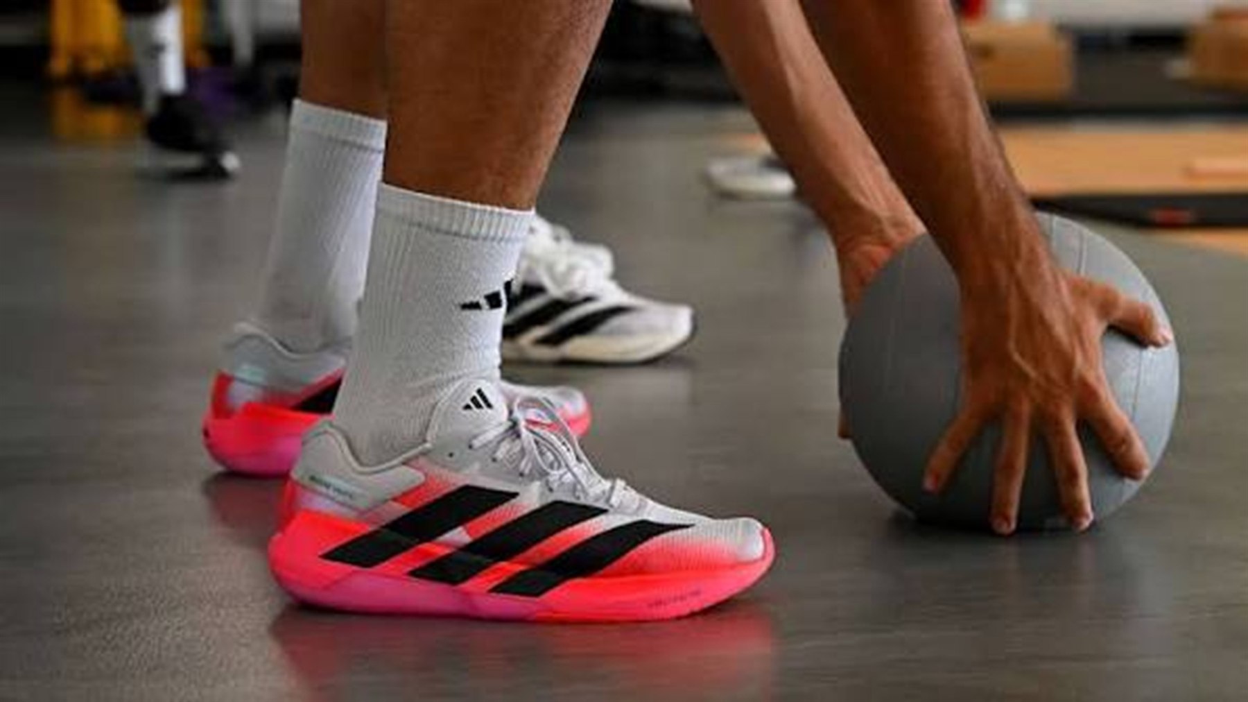 Pés de um atleta calçando o Adizero Dropset Pro branco com entressola coral, apoiado no piso de uma academia ao lado de uma wall ball