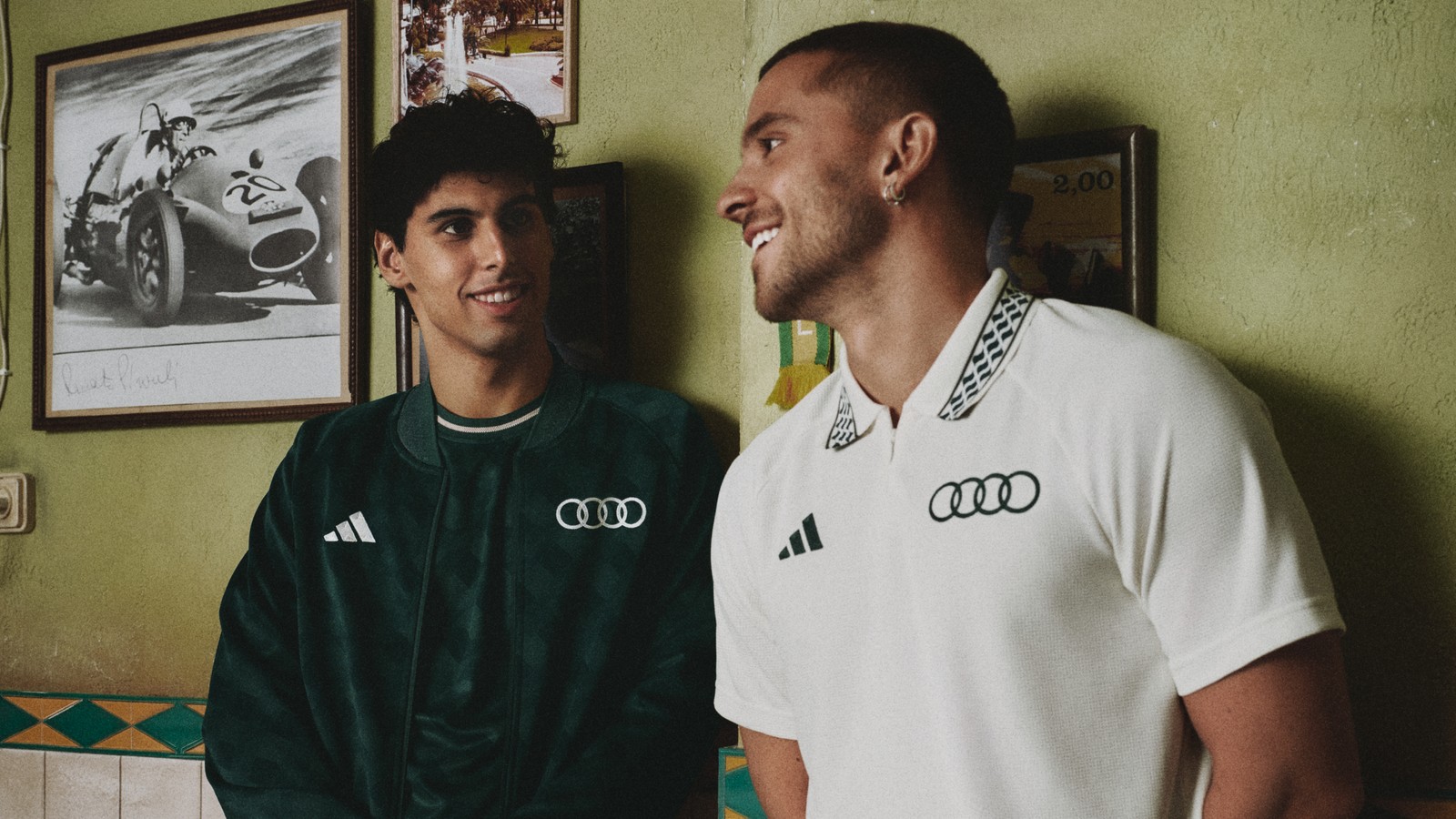 Dois homens conversam num bar de paredes verdes, um com jaqueta verde da Audi e outro com polo branca de gola quadriculada
