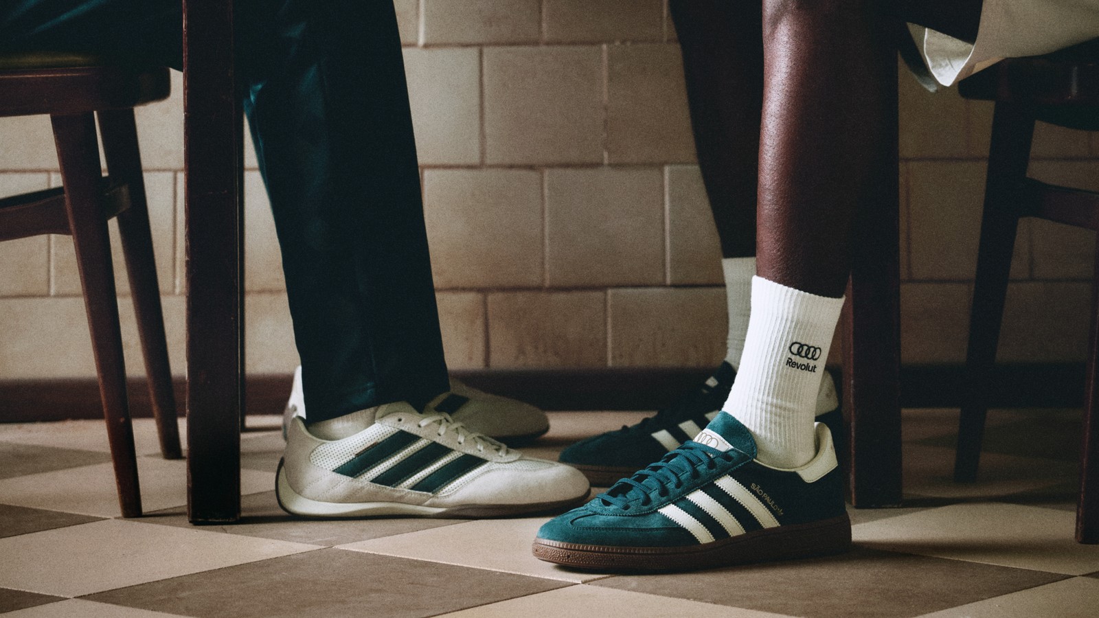 Detalhe dos pés de duas pessoas num piso quadriculado, com tênis adidas brancos e verdes e meia com o logo da Audi