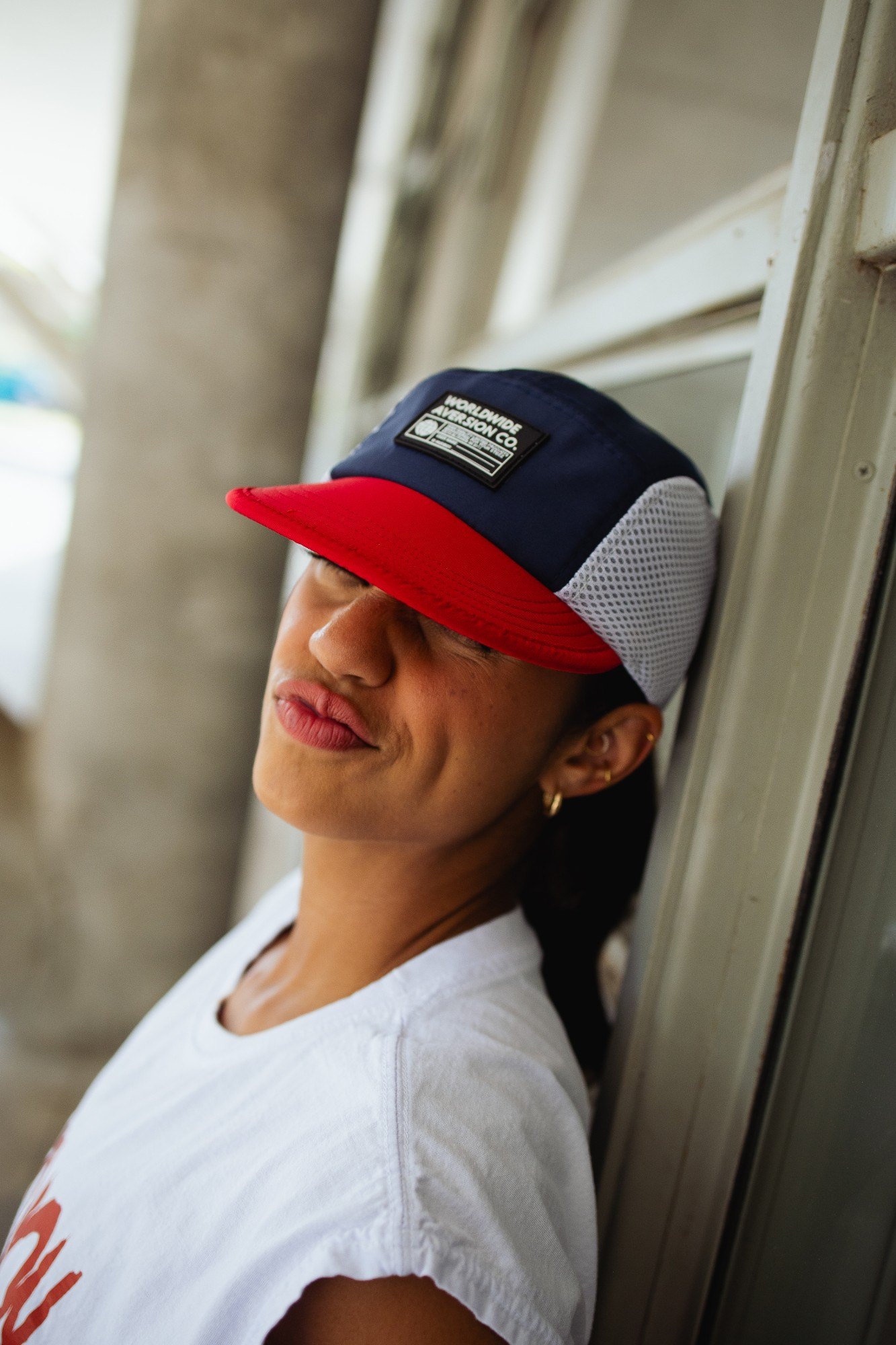 Mulher de olhos fechados encostada numa porta, usando boné five panel azul-marinho com aba vermelha e laterais de tela branca da Aversion