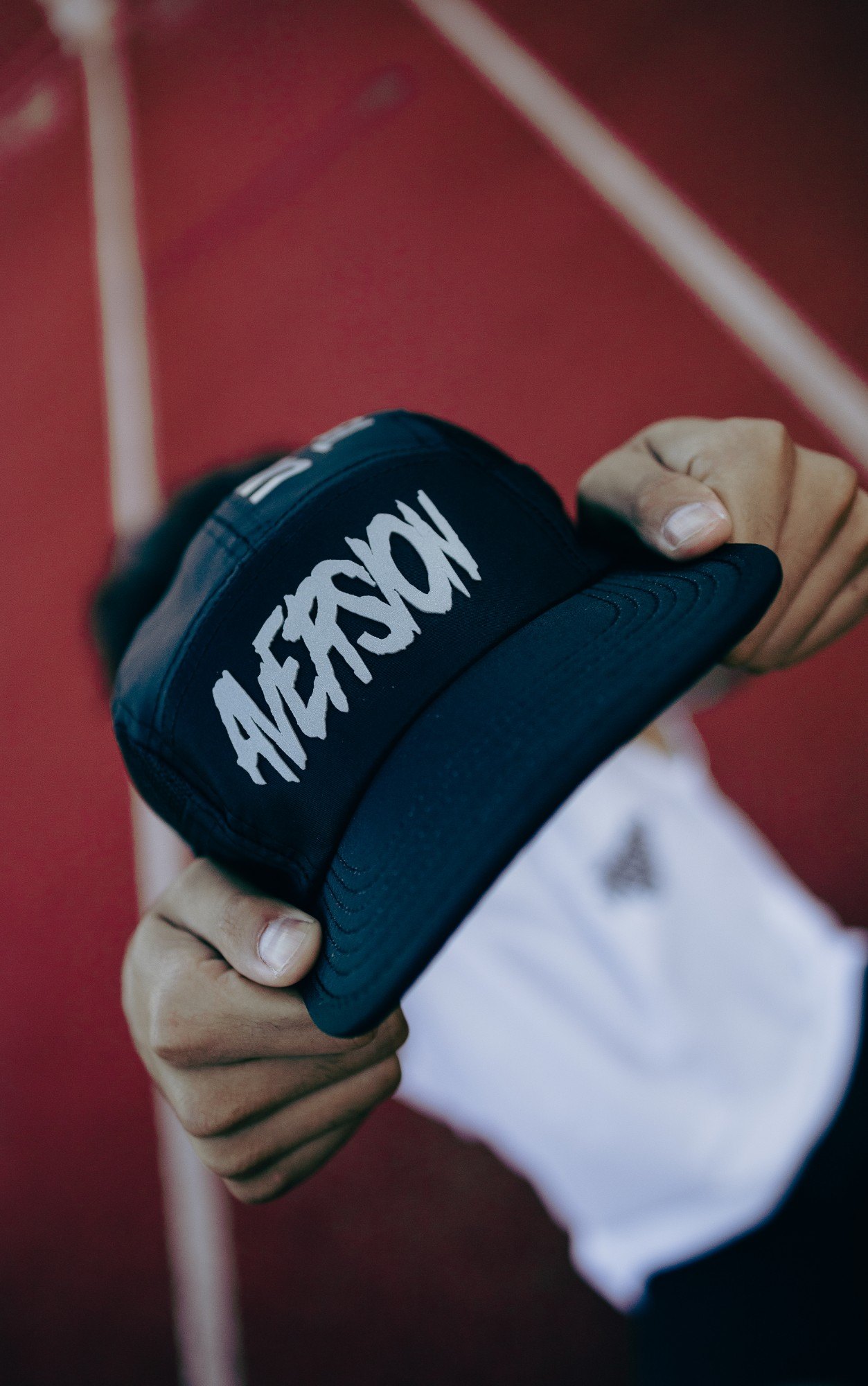 Mãos seguram um boné five panel preto com o nome Aversion em silk refletivo cinza, diante de uma pista vermelha