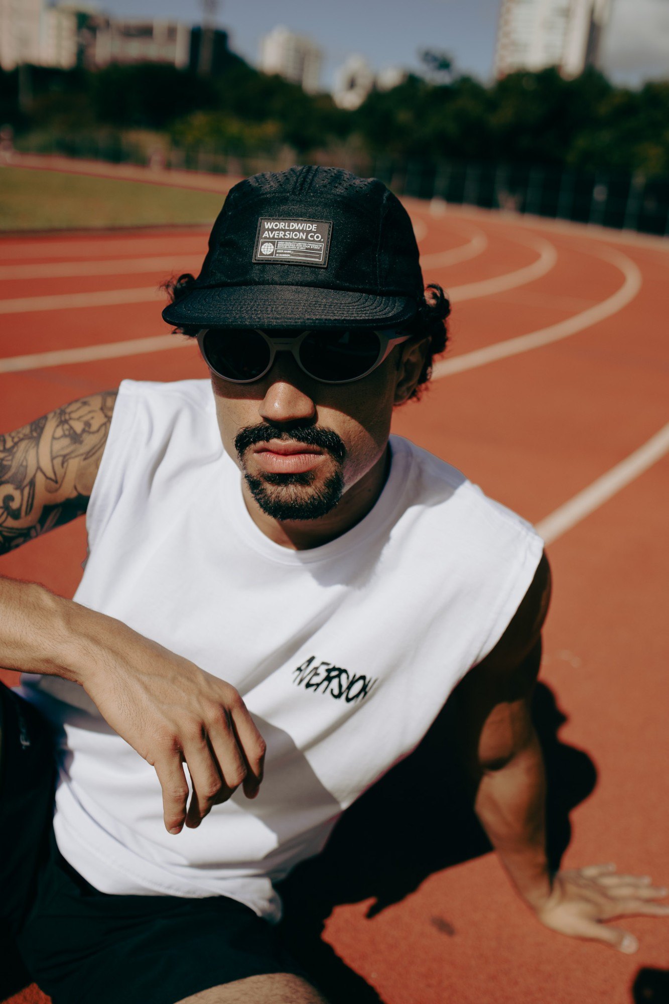Homem de óculos escuros, regata branca e boné five panel preto da Aversion sentado numa pista de atletismo, com prédios ao fundo