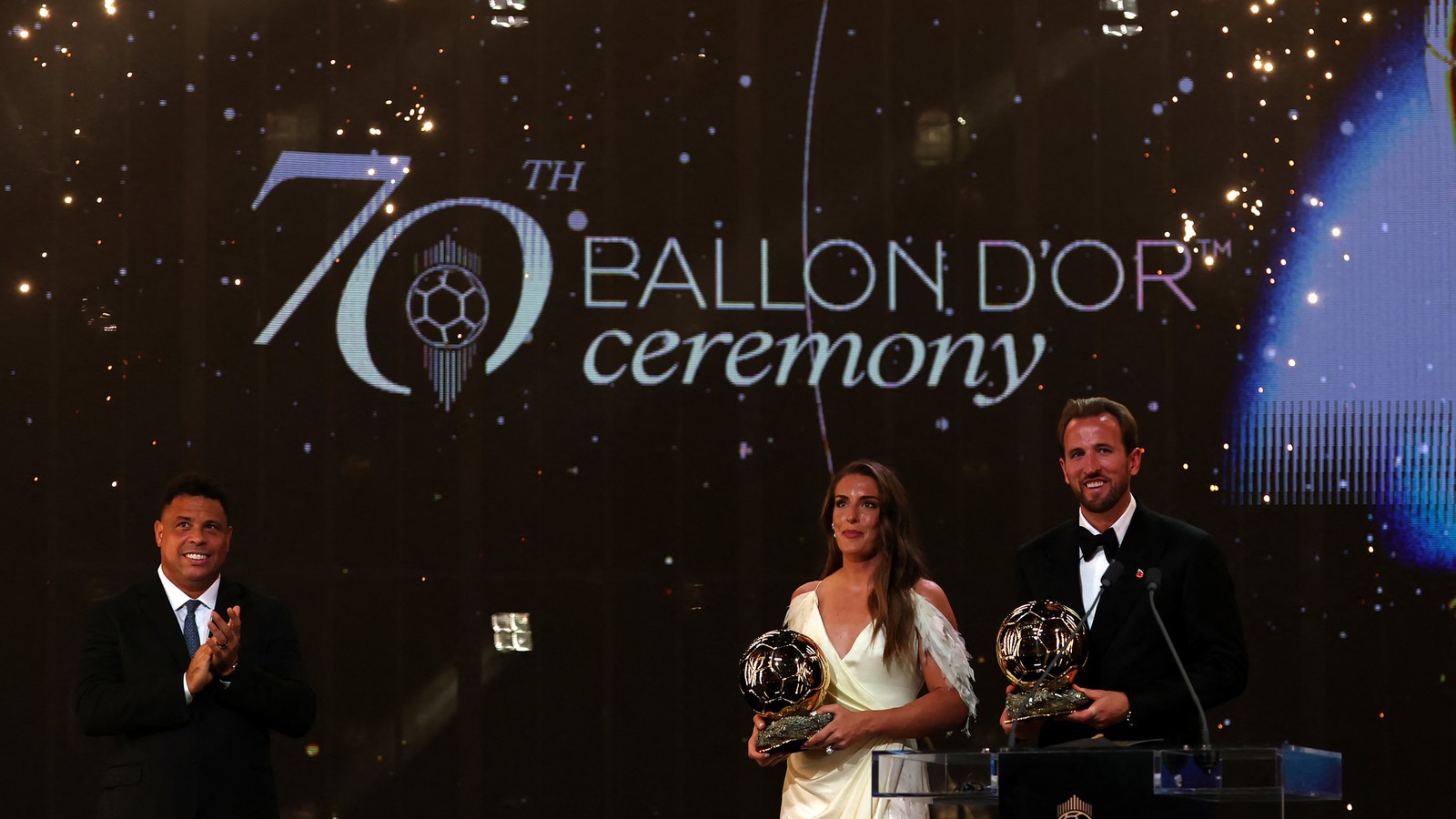 Alexia Putellas e Harry Kane com os troféus no palco da 70ª cerimônia da Bola de Ouro, ao lado de um apresentador que aplaude