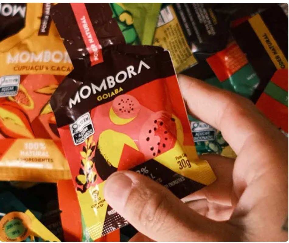 Mão segura um sachê de gel natural Mombora sabor goiaba sobre uma pilha de sachês coloridos