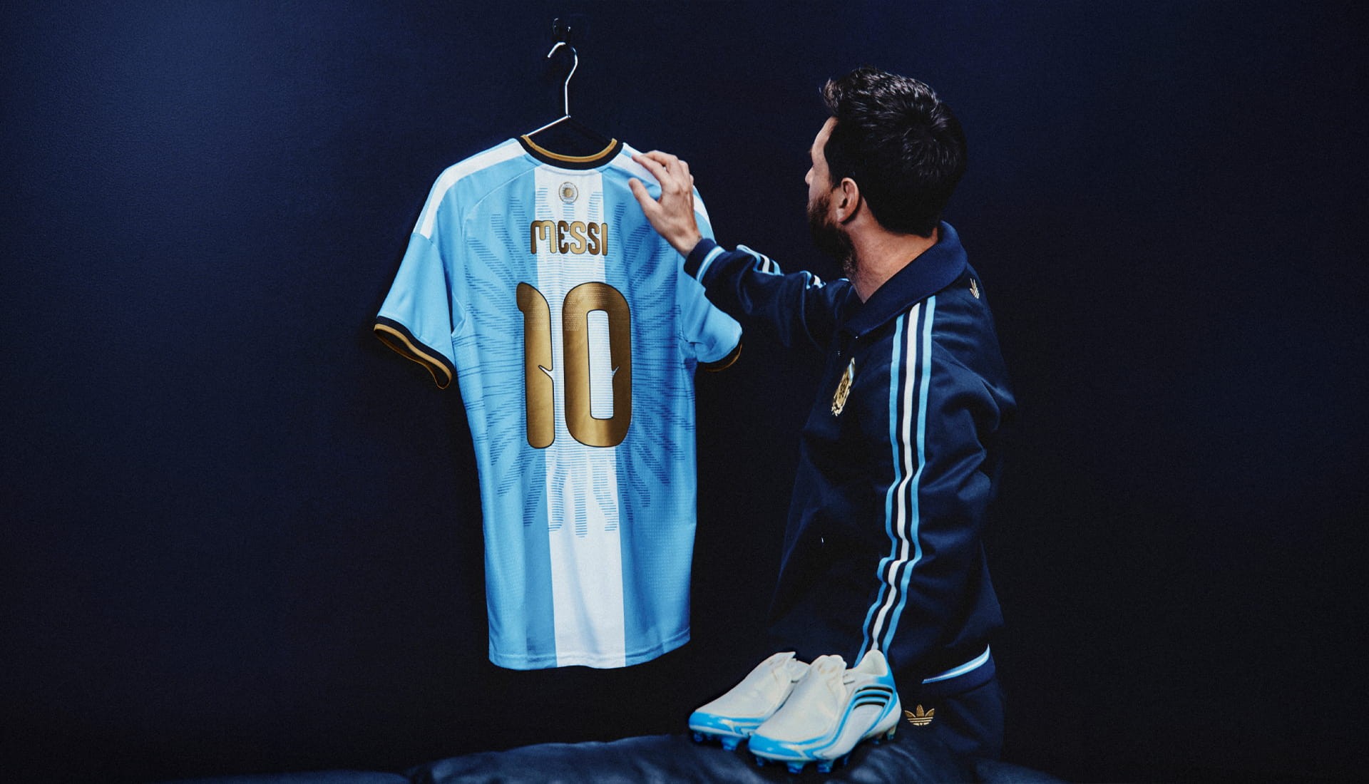 A camisa de despedida de Messi pendurada num cabide, vista de costas, com o nome MESSI e o número 10 em dourado, enquanto o jogador a observa de agasalho azul-marinho