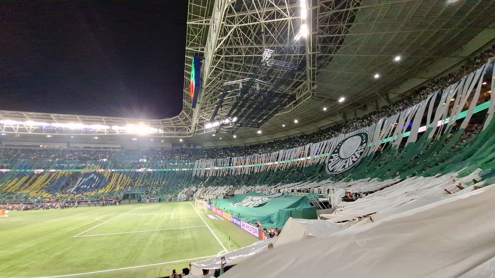 Arquibancada lotada de um estádio brasileiro em noite de jogo, com mosaico da torcida ocupando os anéis superiores