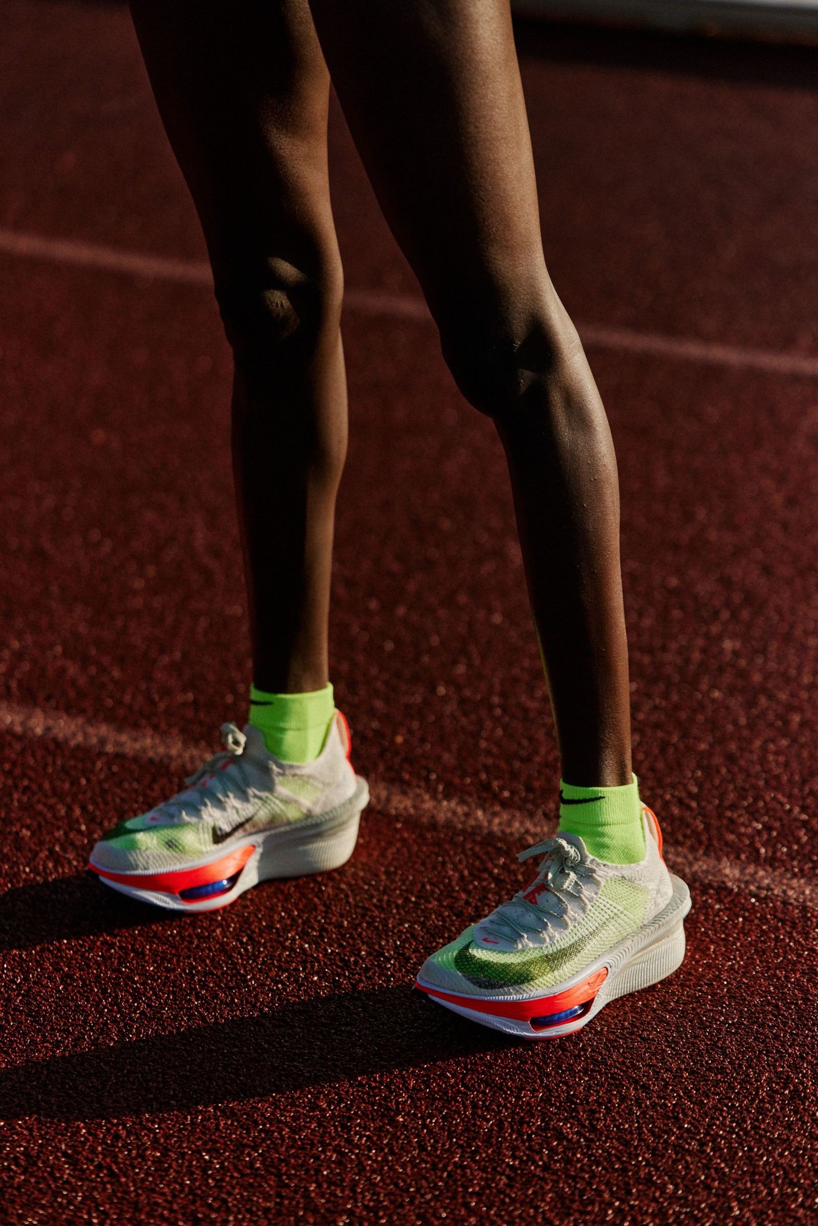 Pernas de Brigid Kosgei em pé na pista de atletismo ao entardecer, calçando o Nike Alphafly 4 branco com meias verde-limão