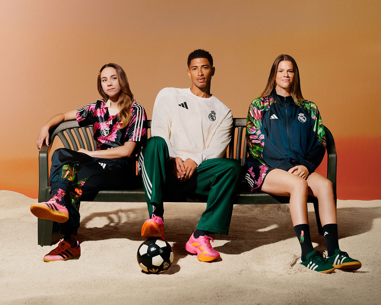 Jude Bellingham sentado num banco na areia, entre duas jogadoras, vestindo moletom creme e calça verde da coleção