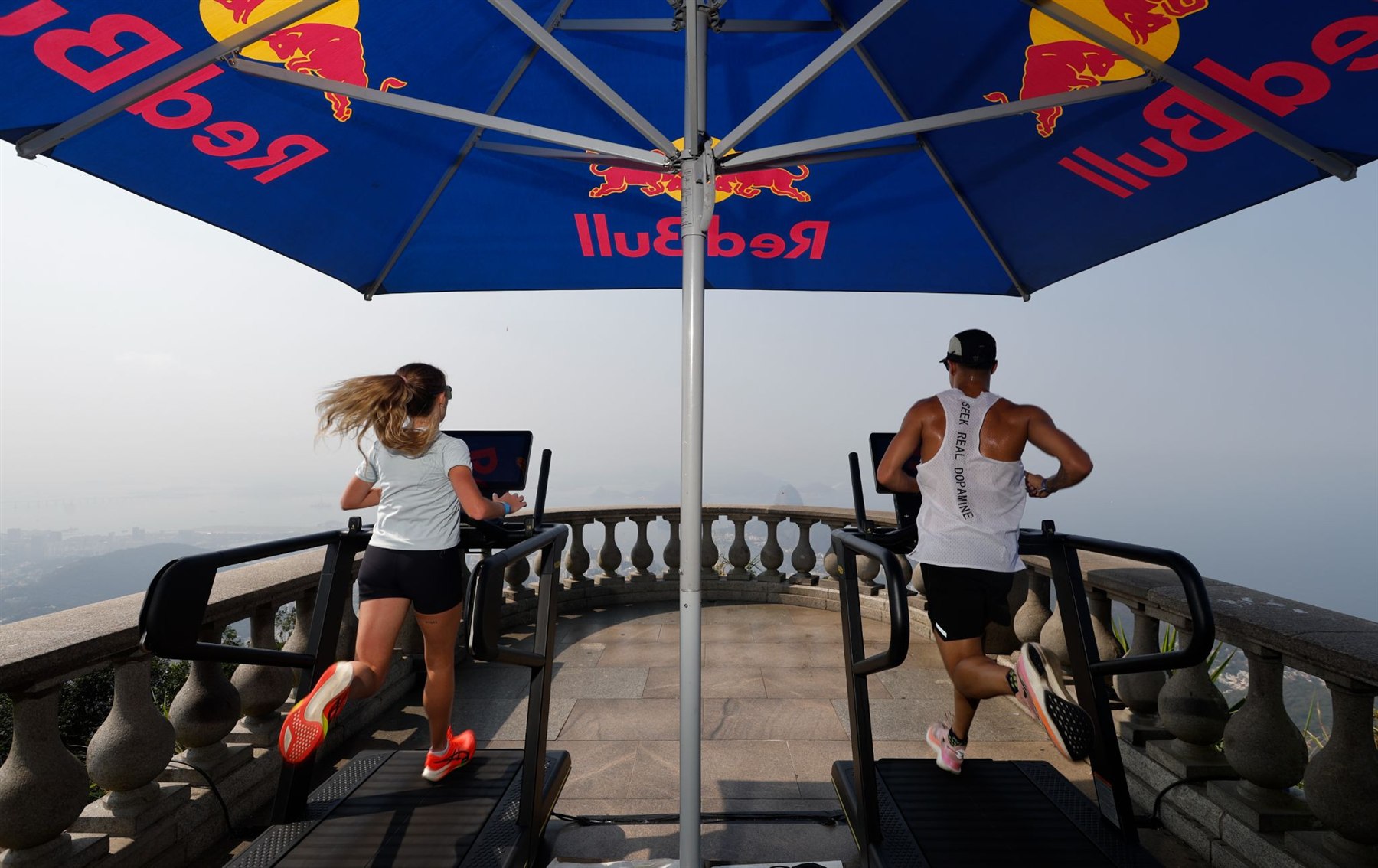 Corredora e corredor lado a lado em esteiras sob um guarda-sol da Red Bull, com a baía do Rio de Janeiro ao fundo
