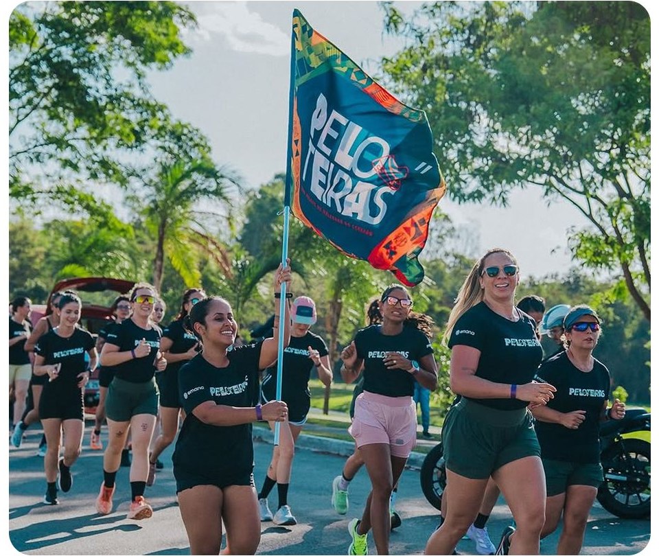 Corredoras do Peloteiras correm em grupo com a bandeira do clube erguida, em dia de sol