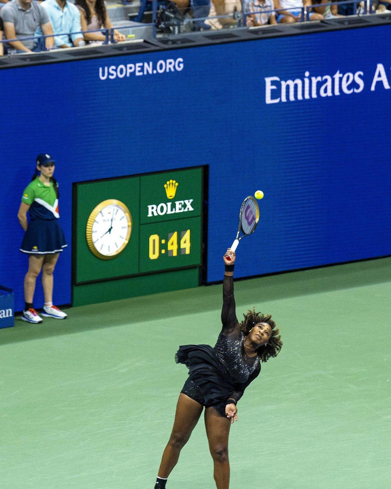 Serena Williams no ar durante um saque no US Open de 2022, com macacão preto brilhante e saia de babado, sob o relógio Rolex da quadra