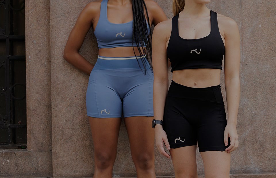 Duas corredoras encostadas numa parede de pedra vestindo o top e o shorts de compressão R.A.W. em azul e preto